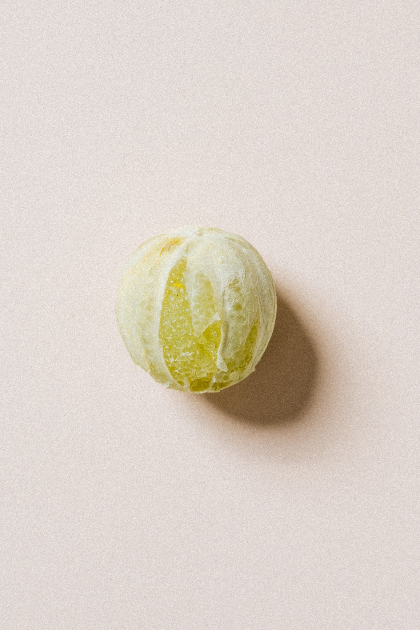 voyage-a-rodrigues A single partially peeled green citrus fruit, likely a mandarin or tangerine, photographed from above on a plain warm beige background with a soft shadow.