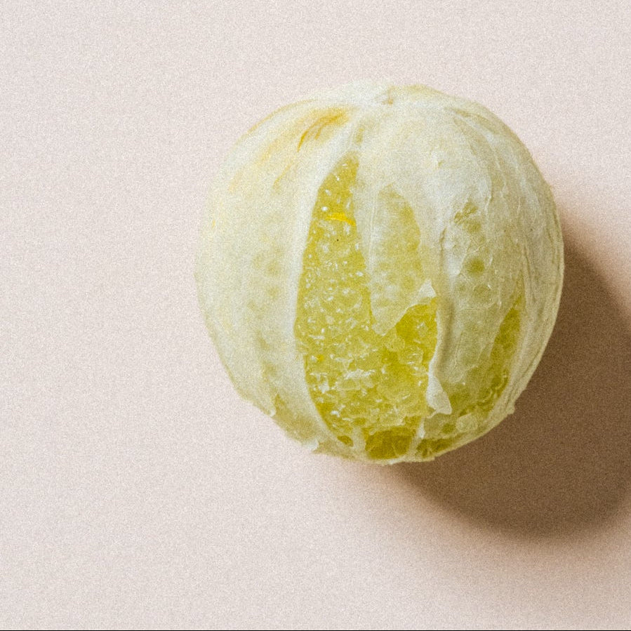 voyage-a-rodrigues A single partially peeled green citrus fruit, likely a mandarin or tangerine, photographed from above on a plain warm beige background with a soft shadow.
