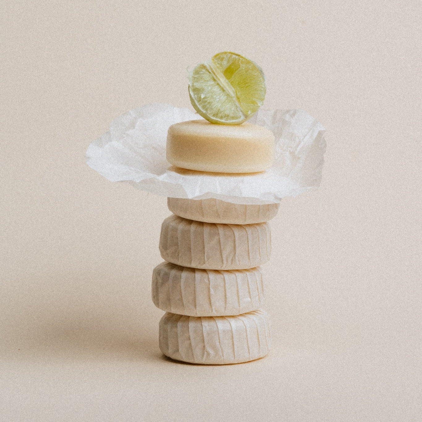 A stack of five round, perfumed soaps wrapped in white paper, topped with a lime wedge, set against a solid beige background.