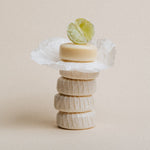 A stack of five round, perfumed soaps wrapped in white paper, topped with a lime wedge, set against a solid beige background.