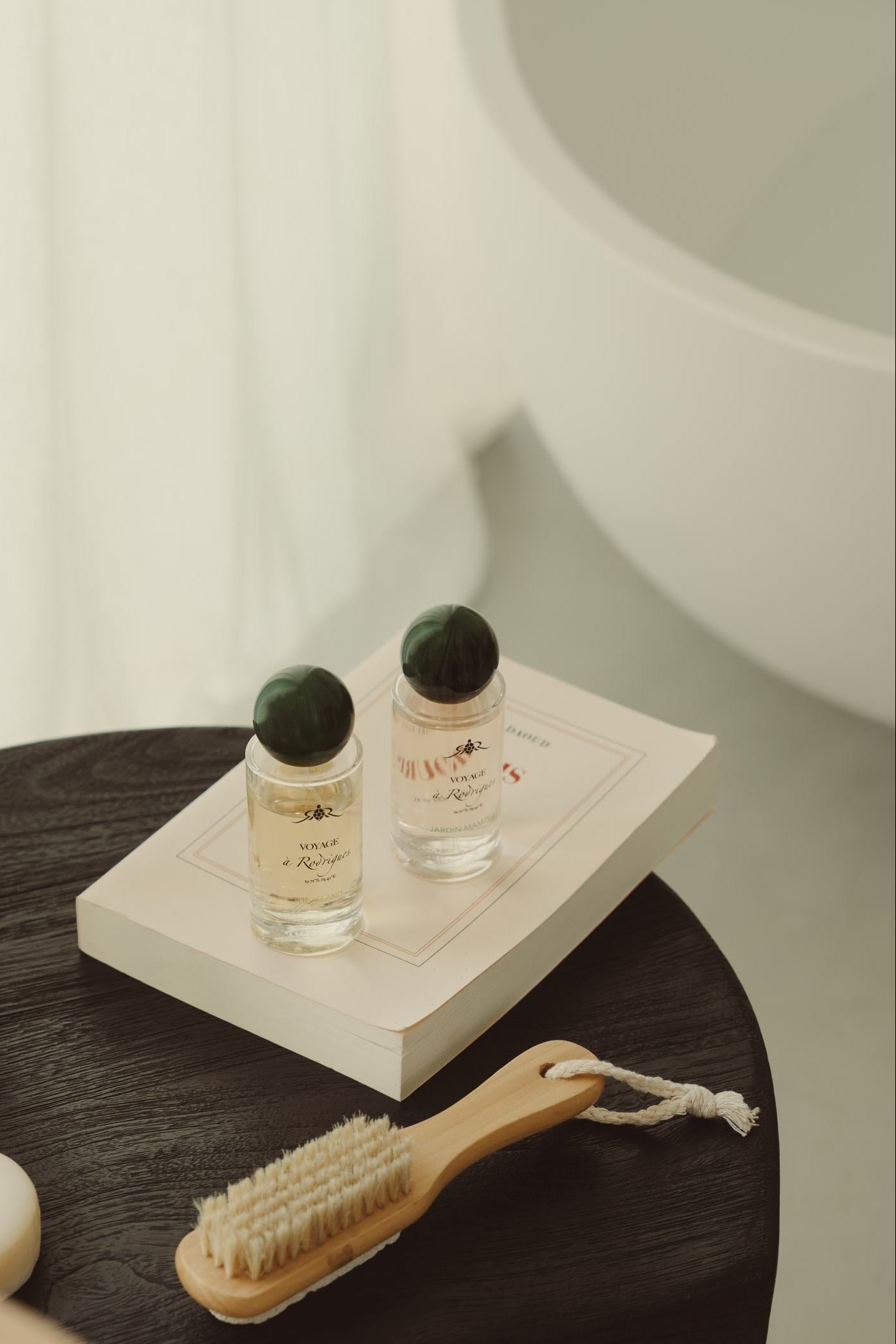 A styled bathroom scene showing several small toiletry bottles and a soap bar arranged on a dark wooden stool beside a wooden brush, with a white tub and soft curtains in the background.